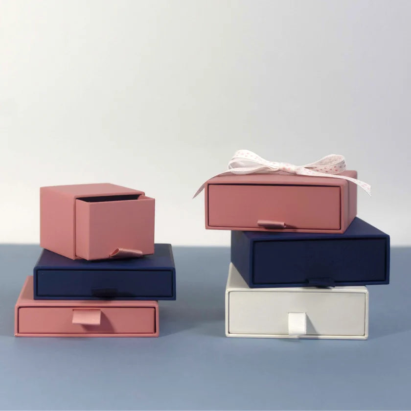 Pastel pink, navy, and cream jewelry boxes are stacked against a pale wall, with a white ribbon draped over the top; the scene feels calm and elegant.