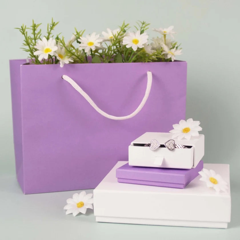 A purple gift bag filled with daisies, next to stacked white and purple gift boxes. A silver bow ring sits on top, conveying a festive and elegant feel.