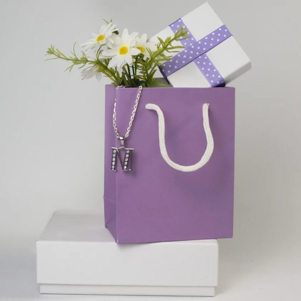 Purple gift bag with a white rope handle, adorned with a jeweled "M" necklace. Inside, daisies and a white gift box wrapped in purple ribbon convey a cheerful, celebratory tone.