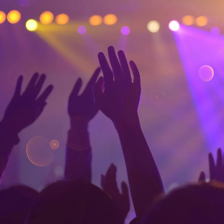 Hands raised in a lively crowd with vibrant purple and yellow stage lights, conveying excitement and energy in a concert atmosphere.