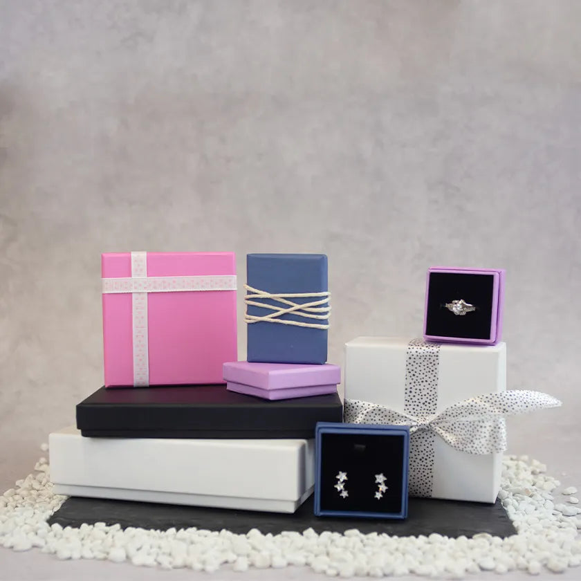 A collection of colourful gift boxes arranged on white pebbles. Includes pink, blue, and white boxes with ribbons.