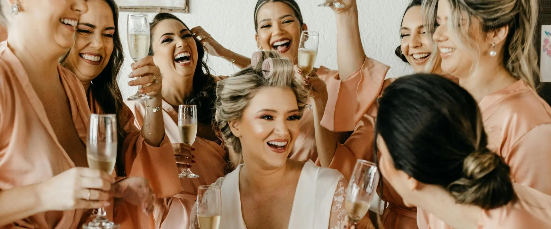 A joyful bride surrounded by bridesmaids in peach robes, all holding champagne glasses. The group is laughing and celebrating in a light-hearted ambiance.
