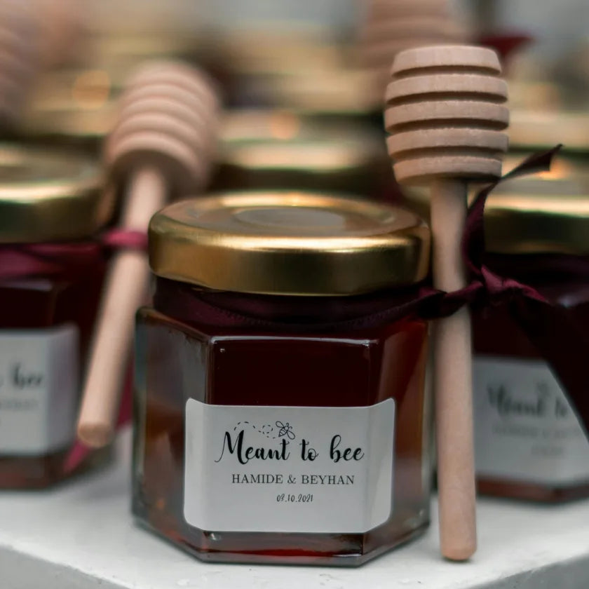 Hexagonal jar of honey with a gold lid and wooden dipper tied by burgundy ribbon. Label reads 