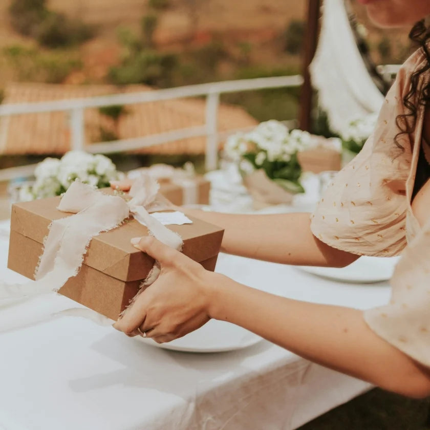 A woman in a light blouse holding a gift-wrapped brown box with a white ribbon at an elegantly set outdoor table adorned with flowers, conveying a celebratory tone.