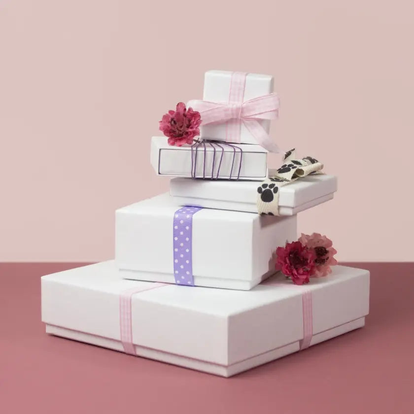 Stack of white gift boxes with pink, purple, and paw print ribbons, adorned with red flowers, on a pink and burgundy background. 